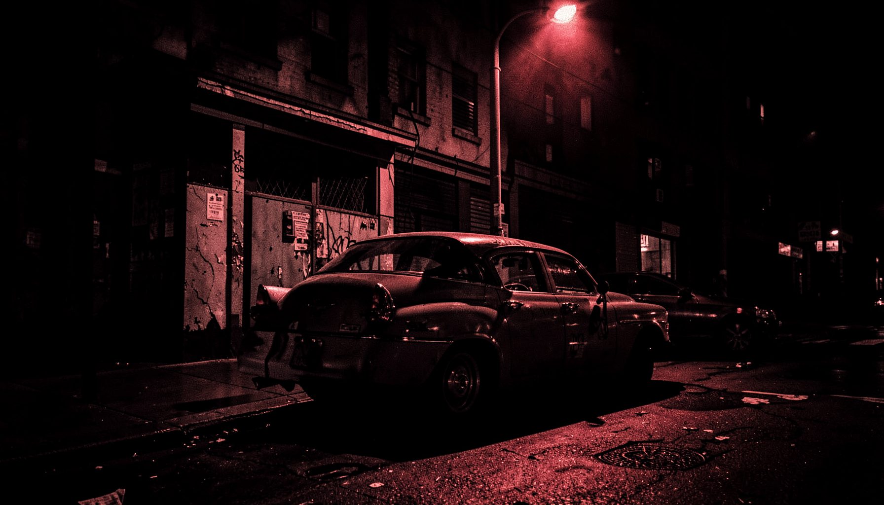 Gritty urban street corner with a vintage car parked under streetlight, deep red and black duotone, tense crime mood