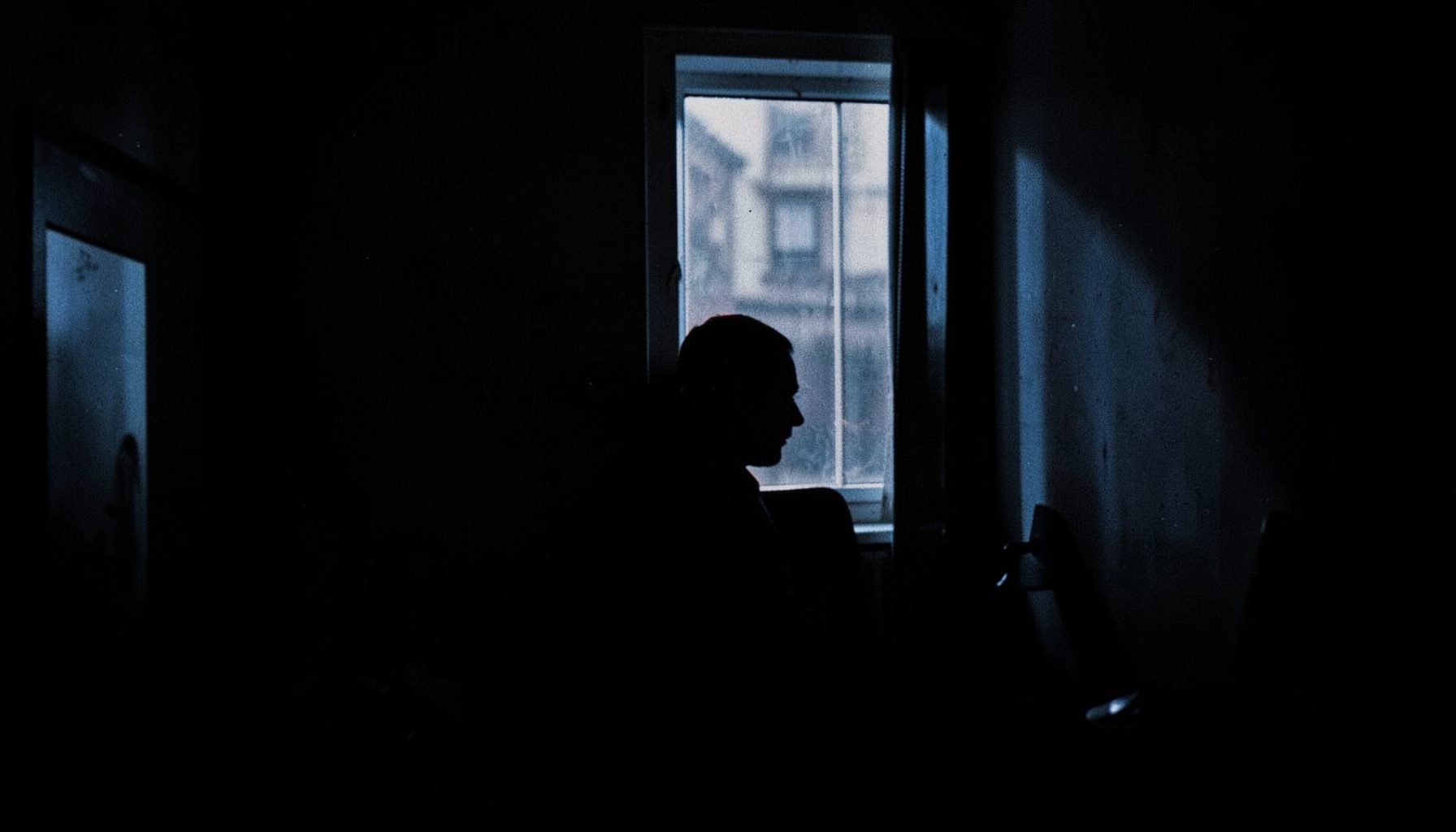 A dimly lit room with a single figure silhouetted against a window, cool blue and shadow tones, tense and contemplative mood
