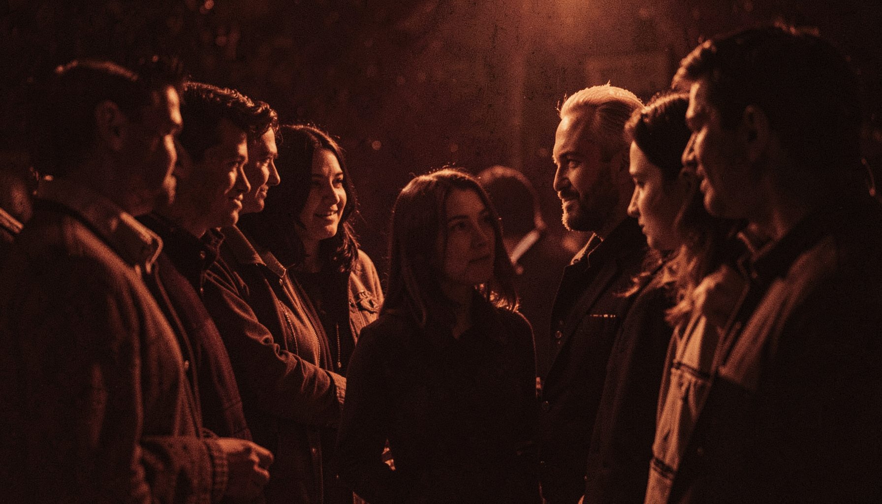 A group of people standing in a circle under warm amber light, their faces partially visible, mysterious and dramatic atmosphere