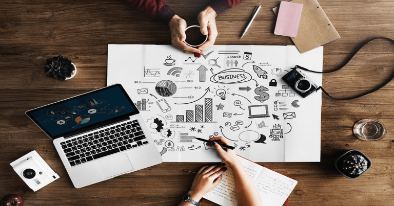 Overhead view of hands writing on a business idea sketch surrounded by laptop, camera, and desk items, warm brown tones