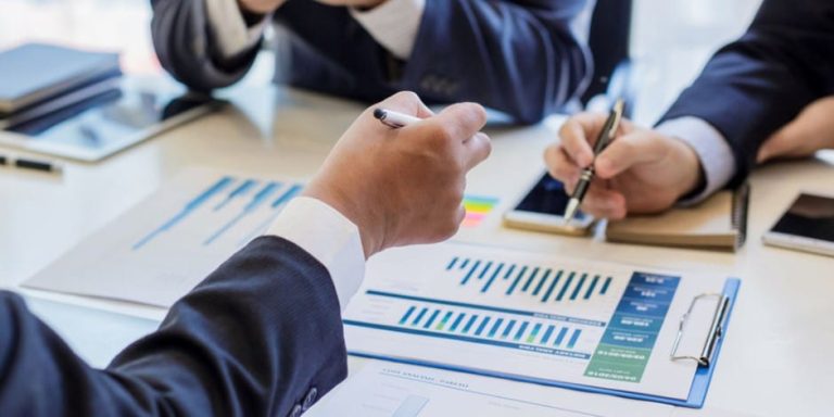 Close-up of business people's hands holding pens over charts and phones on a desk, blue and white tones, professional mood
