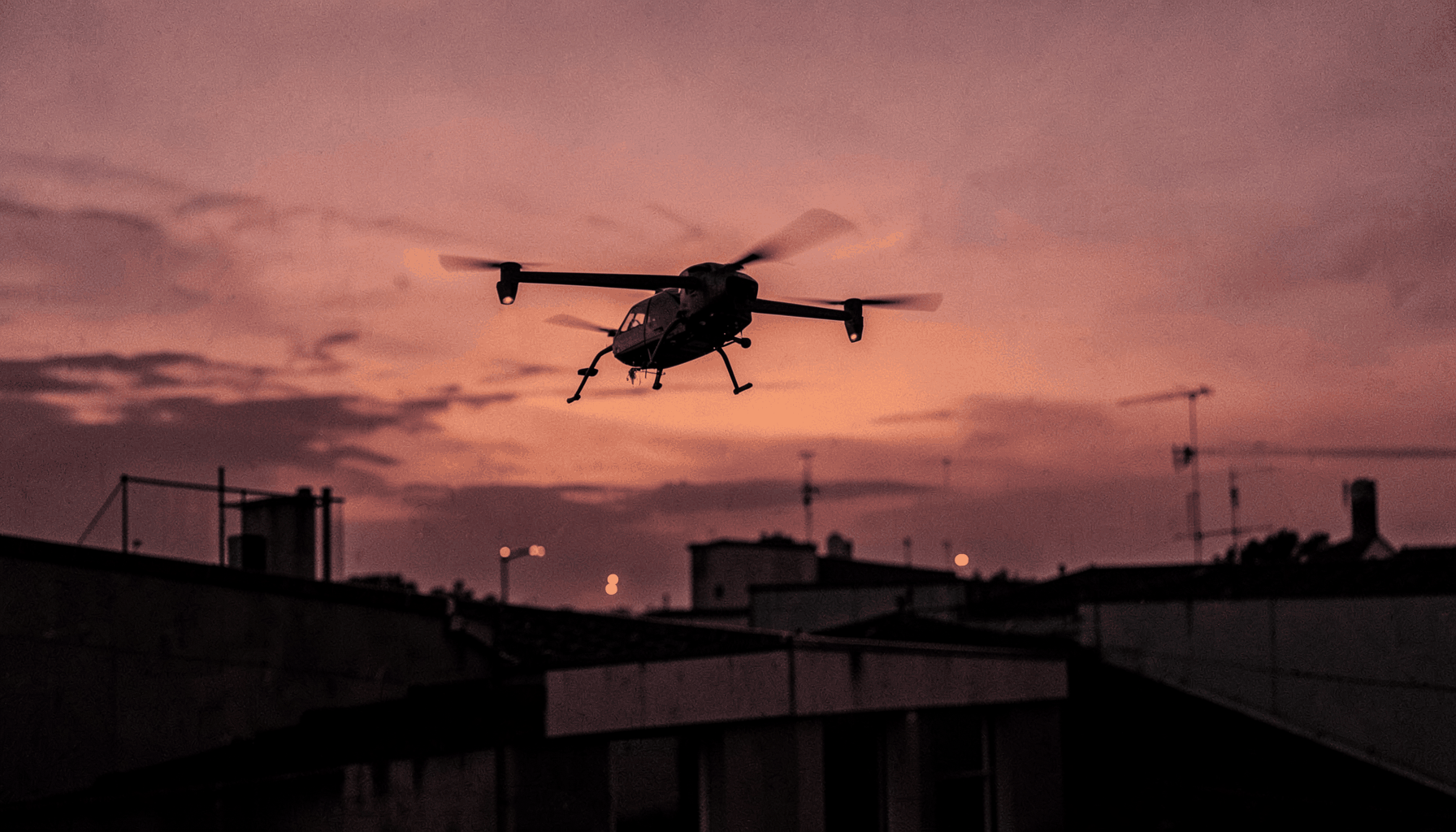 A drone hovering above a rooftop at dusk, orange and purple sky behind it, tense and modern atmosphere